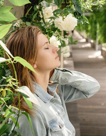Beautiful young blond woman standing in white roses in a garden or park, eyes closedの写真素材