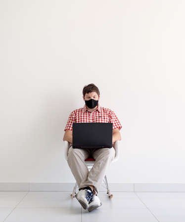 Young caucasian man in plaid shirt wearing a mask, sitting on chair at home or office using laptopの写真素材
