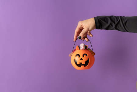 Happy Halloween concept. Scary woman Halloween hand with black nails holding pumpkin full of sweets on purple background with copy spaceの写真素材