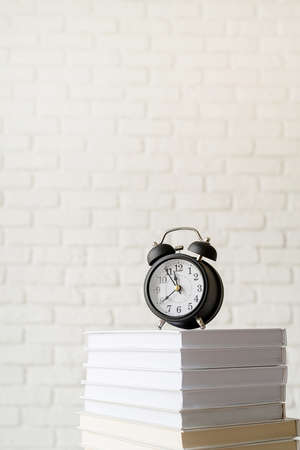 Black alarm clock on stack of books on white brick wall backgroundの写真素材