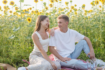 Young teenage couple in white shirts having picnic on sunflower field in sunset. Eating pizza and drinking champagneの写真素材