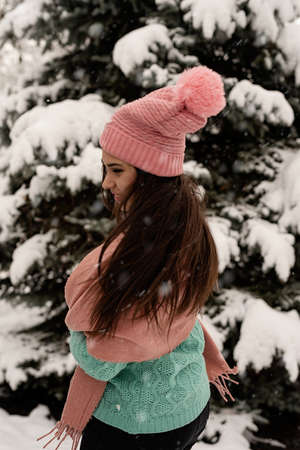 Merry Christmas and happy New Year. Happy Woman in warm winter clothes standing by the big christmas tree outdoors, arms up, snow fallingの写真素材