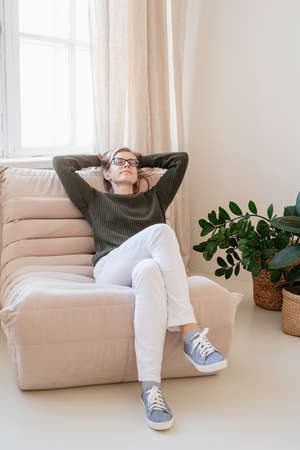 Young thoughtful caucasian woman in green sweater and white jeans sitting on sofa with eyes closed in stylish blue living roomの写真素材