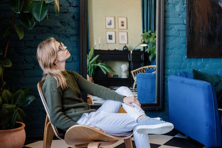 Young thoughtful caucasian woman in green sweater and white jeans sitting on sofa with eyes closed...