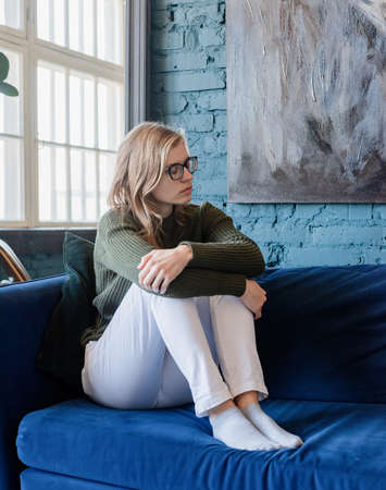 Young thoughtful caucasian woman in green sweater and white jeans sitting on sofa with eyes closed in stylish blue living roomの写真素材