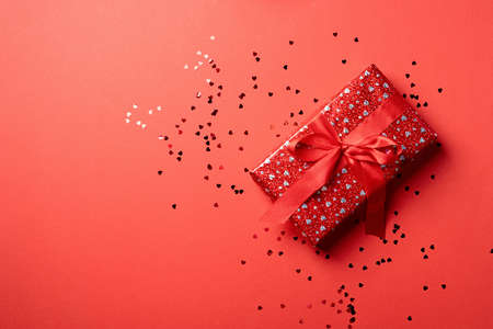 Valentines Day. Red Gift box with beautiful red ribbon with heart shape confetti on red background...