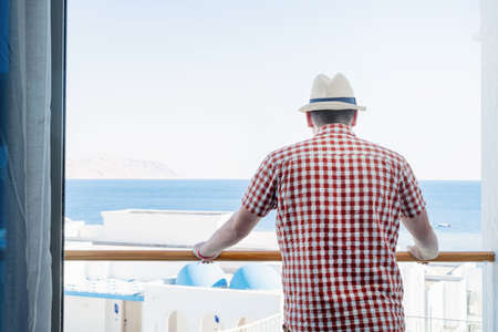 vacation. Handsome Caucasian young man in summer hat standing on balcony of hotel, using mobile phoneの写真素材
