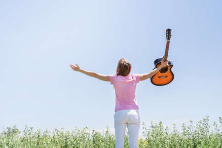 freedom and happiness. Young woman standing in grass fiel raising her hand to the sky, holding guitar, view from behindの写真素材