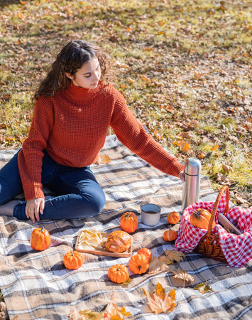 Leisure, free time. Beautiful caucasian woman in red sweater on a picnic outdoors, sitting on a plaid in autumn forestの写真素材