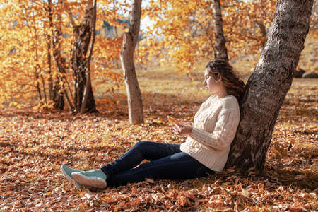 autumn nature. Young thoughtful woman sitting by the tree in autumn forestの写真素材