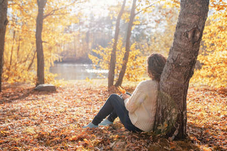 autumn nature. Young thoughtful woman sitting by the tree in autumn forestの写真素材