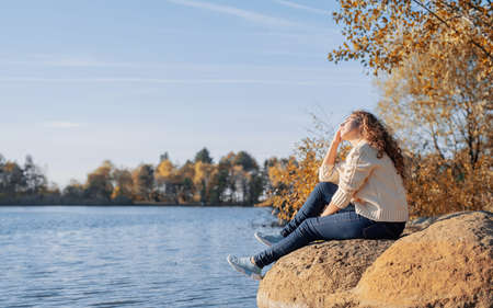freedom concept. Thoughtful romantic woman sitting on rocks on the river bank in sunset in autumn day, copy space.の写真素材