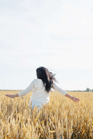 Mid adult woman in white dress walking and dancing across golden field holding heap of rye lit by...