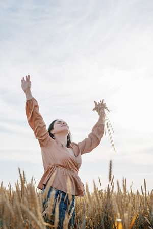 Mid adult woman in beige shirt walking across golden field holding heap of rye lit by sunset light, copy spaceの写真素材