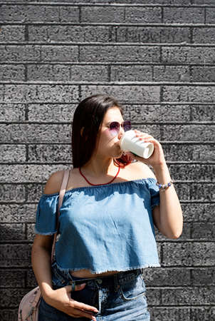 stylish young woman wearing jeans shirt, sunglasses and red jewelry at street, white wall...