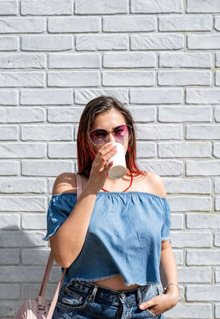 coffee to go. stylish young woman wearing jeans shirt, sunglasses and bag, drinking coffee to go at street, black brick wall backgroundの写真素材