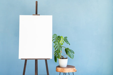 Wooden easel with blank canvas with monstera plant on light blue wall background for mockup design. Space for textの写真素材