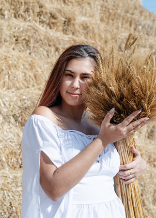 Picnic country side. Young woman sitting on haystack in harvested field holding wheat bouquet