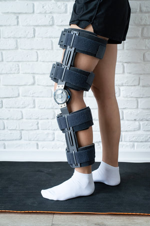 Woman after car accident in an orthosis standing on sport mat. Female wearing knee orthosis or knee support brace and crutches after surgery on leg front viewの写真素材