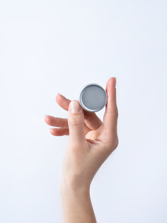 Woman hand holding blank round tin container for lip balm or cream on light gray background....