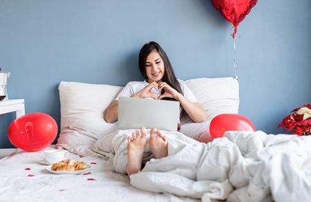 Valentine's day. Young happy brunette woman sitting in the bed with red heart shaped balloons...