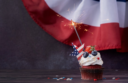 Fourth of july celebration. Sweet cupcake with blueberries and strawberry and sparkler over USA flag backgroundの写真素材