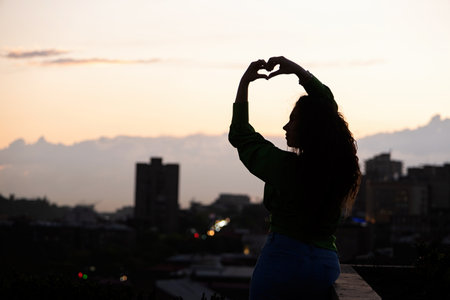 silhouette of beautiful woman on rooftop at urban sunset, making heart shape with handsの写真素材