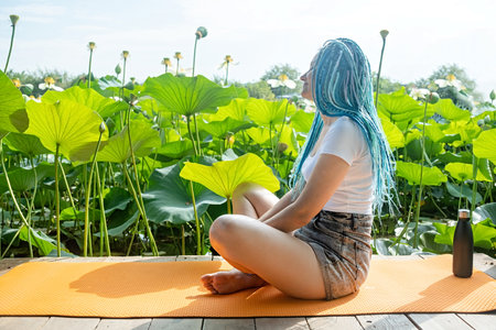 young beautiful woman with blue afro locks resting on yoga mat on wooden pierce on lotus lake enjoying natureの写真素材