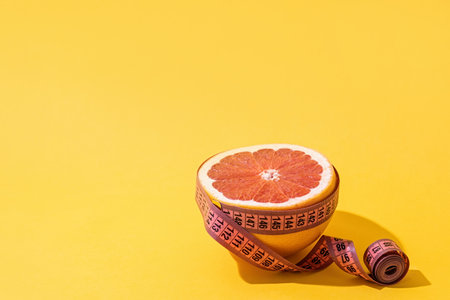 Slimming, body shaping, skin care. Anti-cellulite creative concept. Fruit orange is wrapped with measuring tape on yellow background.の写真素材