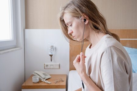 thoughtful woman in earphones sitting on bed in hotel room using mobile phoneの写真素材