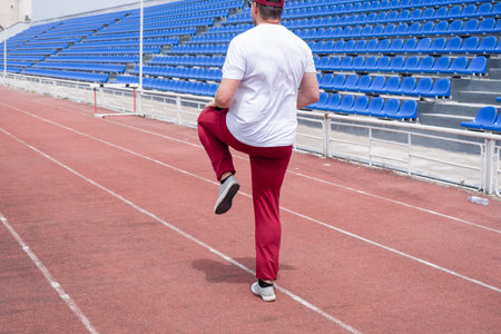 Athletic bearded man wearing red pants and cap warms up before workout at the stadium. Space for...