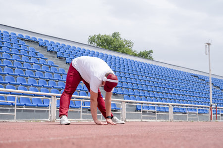 Athletic bearded man wearing red pants and cap warms up before workout at the stadium. Space for textの写真素材