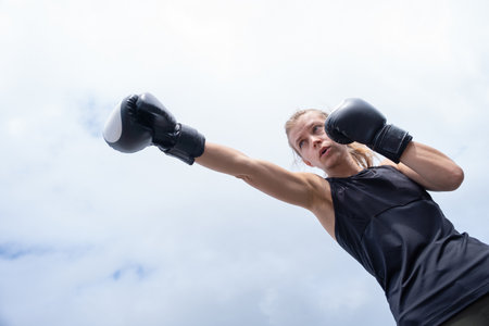 young female boxer wearing sport clothes and gloves standing smiling on sky background, showing arms musclesの写真素材