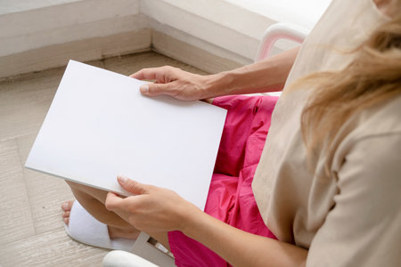 reading on vacation. unrecognizable woman wearing pink shorts and beige tshirt shitting on the hotel balcony holding blank magazine for mockup designの写真素材