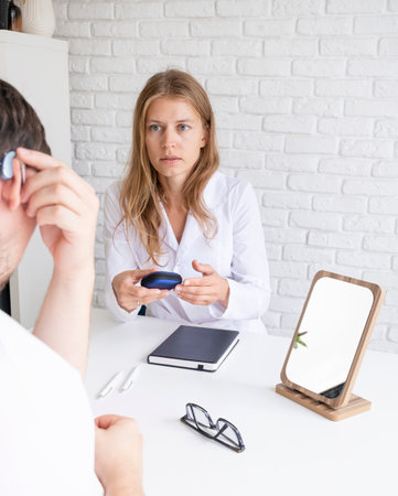 young male patient testing hearing aid in clinic exam room, young female doctor helping