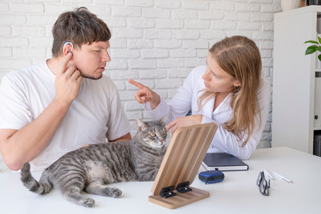 young male patient testing hearing aid in clinic exam room, young female doctor helping