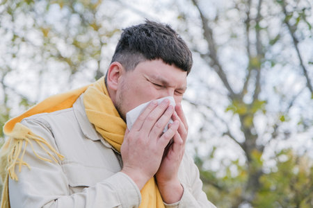 young man wearing yellow scarf enjoying autumn days using tissue to wipe nose, seasonal allergy