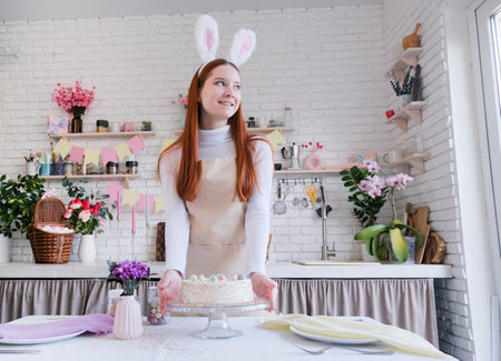 Happy Easter. young woman wearing bunny ears holding Easter cake in the kitchen, getting table ready for holidayの写真素材