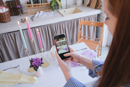 Happy Easter. Young millennial woman in blue shirt coloring preparing in the kitchen, taking photo on mobileの写真素材