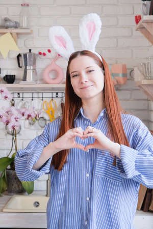 Caucasian woman in bunny ears standing by serving table for Easter dinner, making heart shape with...