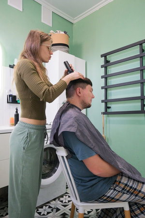 young caucasian woman cutting man hair in bathroom at home using electric razor machineの写真素材
