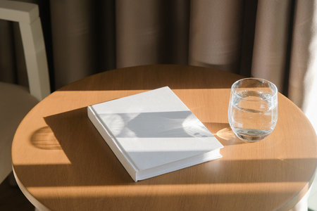 book cover mockup design. blank book mockup on round wooden coffee table with glass of water in the...