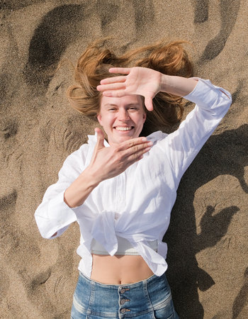 top view of happy young woman lying on sand enjoying vacation arms outstretched, making frame sign with handsの写真素材