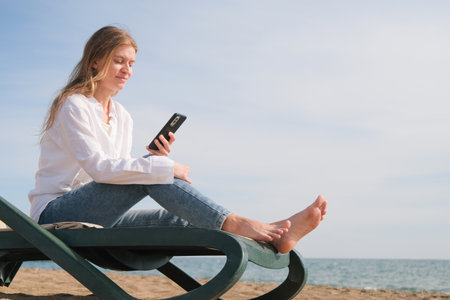 young woman sits in the chaise lounge on the edge of the sea using smart phoneの写真素材
