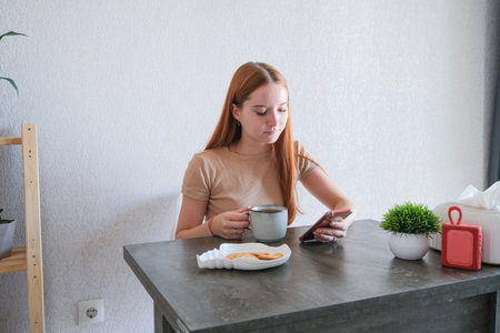young redhead woman drinking tea and using smartphone in the kitchen