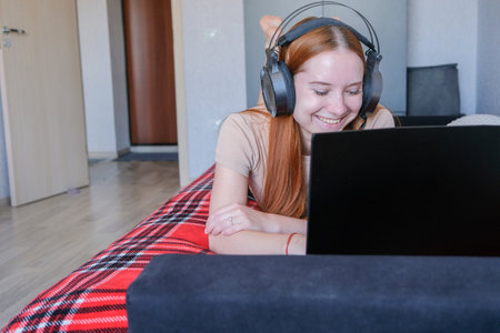 Young redhead woman wearing headphones working on laptop lying on the bed. Female working from home,...