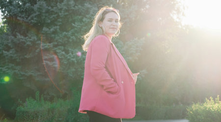 Happy plus size Caucasian woman in pink jacket and shoes walking outdoors, lit from behindの写真素材