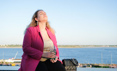 portrait of beautiful caucasian plus size woman in pink jacket and shoes sitting on the embankment...