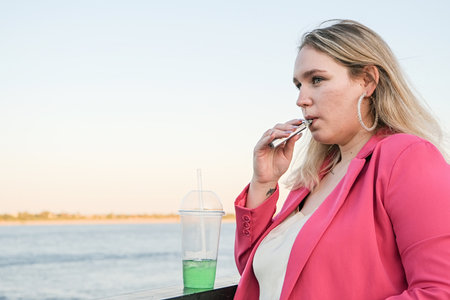 Plus size teenager girl in casual clothes smoking an electronic cigarette on the river embankment outdoors in summer day. Bad habit harmful to health. Vaping activity.の写真素材