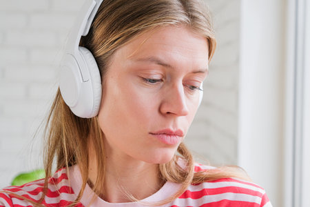 young millennial woman listening to the music at home standing next to the window feeling sadの写真素材